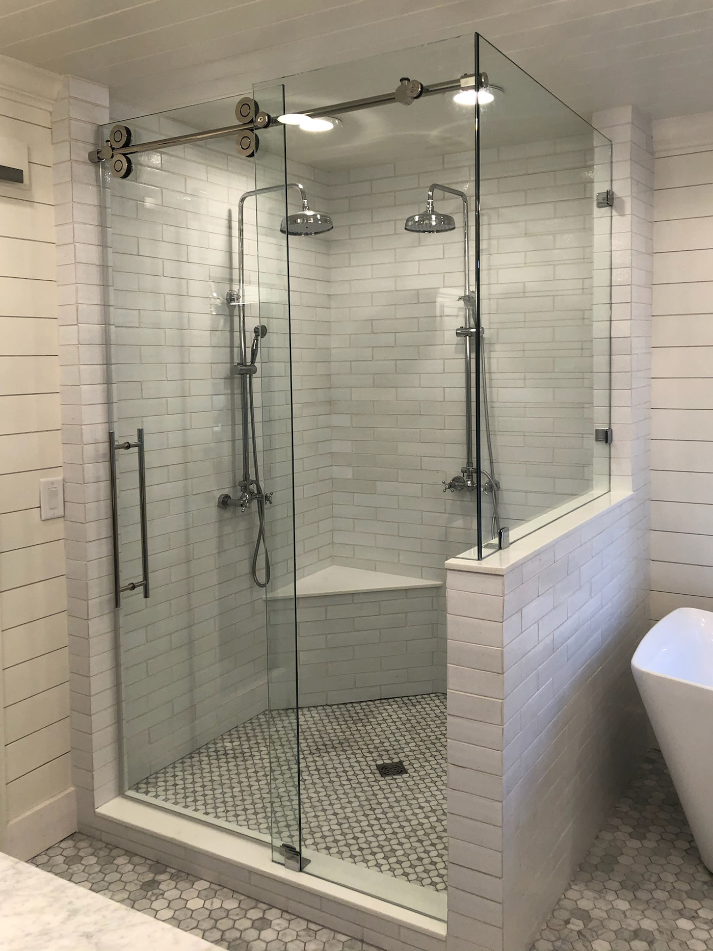 Modern bathroom corner shower with glass sliding door, dual shower heads, built-in bench, and white subway tile walls.