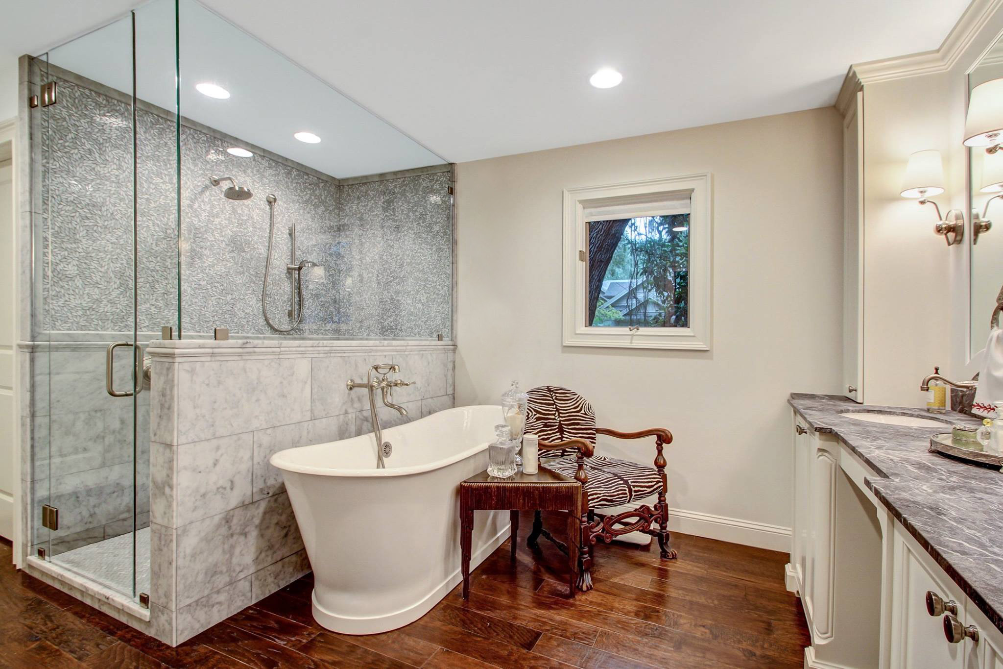 Elegant bathroom with a white freestanding tub, glass-enclosed shower with marble walls, wooden floor, and a zebra-patterned wooden chair beside a small table.